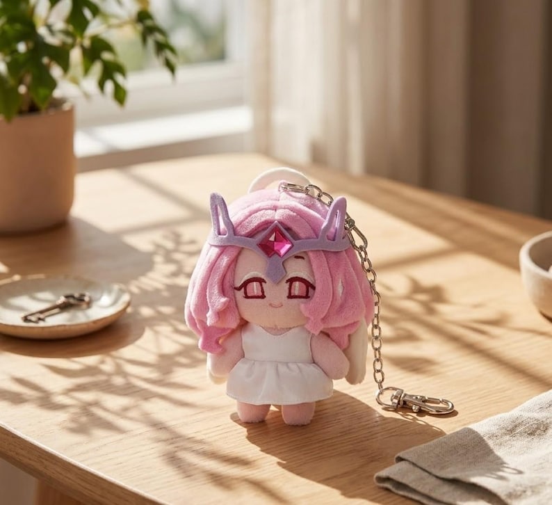 May include: A plush doll with pink hair, a purple crown, and a white dress. The doll has a keychain attached. The doll is on a wooden table.