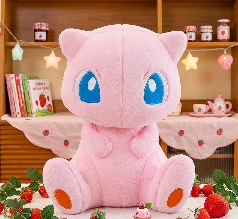 May include: A large, pink plush toy of a cartoon character with large blue eyes and orange accents on its feet. The plush is sitting in front of a shelf with books, jars, and decorative lights. Strawberries and strawberry decorations are in the foreground.