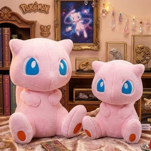 May include: Two plush pink Mew Pok&eacute;mon figures with large blue eyes and orange accents. The figures are sitting on a patterned surface, with a framed picture of Mew in the background. The word "Pok&eacute;mon" is visible.