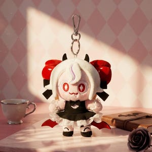 May include: A plush doll with a white wig, red eyes, and small black horns. The doll wears a black dress with a white ruffled skirt and red accents. It has a silver keychain attached. A teacup, book, and rose are in the background.