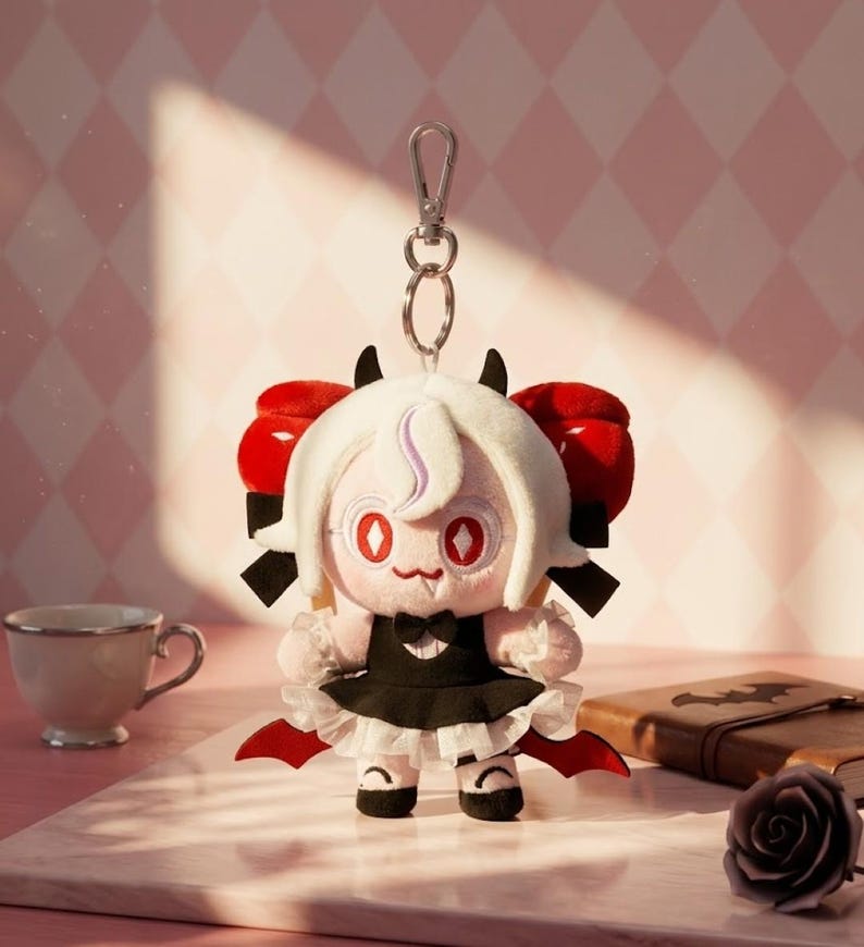 May include: A plush doll with a white wig, red eyes, and small black horns. The doll wears a black dress with a white ruffled skirt and red accents. It has a silver keychain attached. A teacup, book, and rose are in the background.