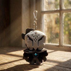 May include: A plush keychain featuring a character with a silver mask and black outfit. The keychain has a silver clasp and ring. The character has a hat and a blue gem on the chest. The background is a wooden surface and a window.