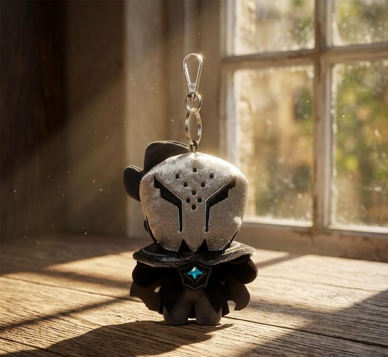 May include: A plush keychain featuring a character with a silver mask and black outfit. The keychain has a silver clasp and ring. The character has a hat and a blue gem on the chest. The background is a wooden surface and a window.