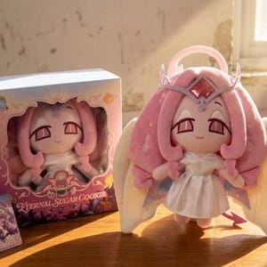 May include: A plush doll with pink hair, a crown, and angel wings. The doll is wearing a white dress and has a small pink halo. The doll is standing next to a box with the same doll inside. The box and doll are labeled "Eternal Sugar Cookie".