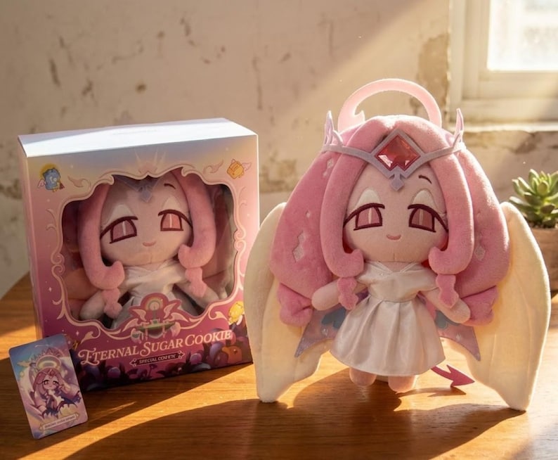 May include: A plush doll with pink hair, a crown, and angel wings. The doll is wearing a white dress and has a small pink halo. The doll is standing next to a box with the same doll inside. The box and doll are labeled "Eternal Sugar Cookie".