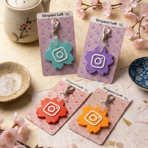 May include: Four colorful keychains in the shape of a flower with an Instagram logo in the center. The keychains are teal, purple, red, and orange. Each keychain is attached to a silver clasp and displayed on a patterned card with the text "Origami Loft".