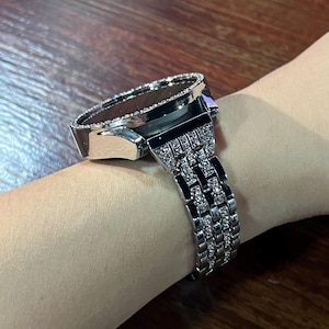 May include: A silver-toned wristwatch featuring a round face surrounded by a border of small clear stones. The metal link band is also embellished with rows of small clear stones, creating a reflective and textured appearance on the wrist.