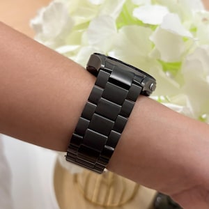 May include: A dark gray watch with a matching metal band is shown on a wrist. The watch band is made of rectangular links. The watch face is visible, and the overall design is sleek and modern. The watch is a stylish accessory.
