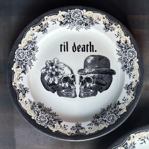 May include: A white ceramic plate with a black and cream floral border. The plate features two skull illustrations, one with a floral headpiece and the other wearing a hat. The text "til death." is printed on the plate.