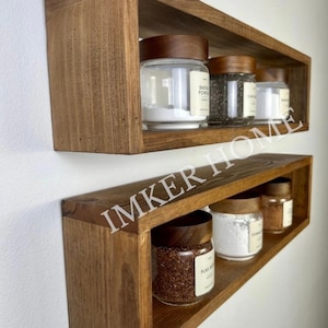 May include: Two wooden shelves with a rectangular frame, mounted on a white wall. The shelves are filled with glass jars with wooden lids, containing various spices and powders. The jars are labeled with text, including "Baking Powder" and "Flax Seed".