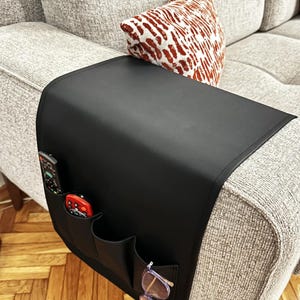 Puede incluir: Organizador de reposabrazos de sofá de cuero negro con bolsillos para mandos a distancia y gafas. El organizador está sujeto al reposabrazos de un sofá gris.