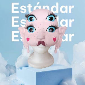 May include: A pink face mask with blue eyes, pink hearts, and a brown mustache. The mask is on a white mannequin head and is sitting on a light blue cube.