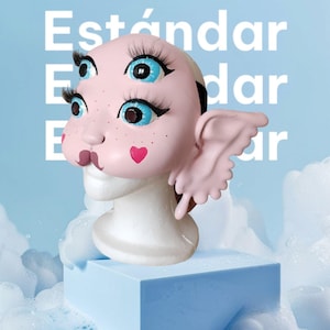 May include: A pink and white doll head with blue eyes and pink wings. The doll head is on a white stand and has a pink heart on its cheek. The doll head is in front of a blue background with the word "Est&aacute;ndar" written in white.