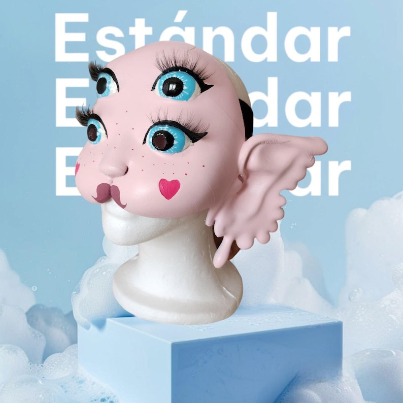 May include: A pink and white doll head with blue eyes and pink wings. The doll head is on a white stand and has a pink heart on its cheek. The doll head is in front of a blue background with the word "Est&aacute;ndar" written in white.