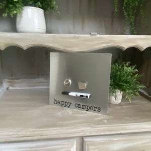 May include: A silver metal magnetic board with the text "happy campers" written in black. The board has a white dry erase marker and a small silver object attached to it.
