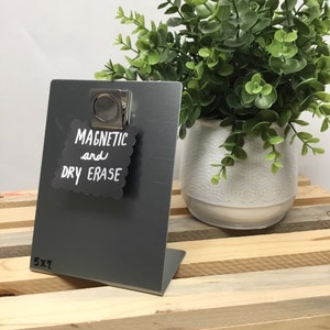 May include: A gray metal dry erase board with a magnetic clip. The board is 5x7 inches and has the words "Magnetic and Dry Erase" written on it in white.