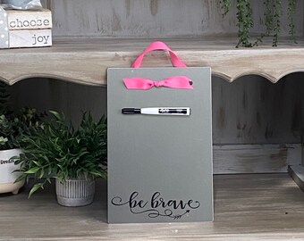 May include: A grey rectangular chalkboard with a pink ribbon handle and bow. A black and white marker is attached. The words "be brave" are written in cursive script with an arrow design.