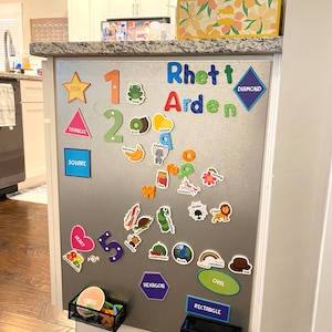 May include: A silver magnetic board with colorful magnets featuring shapes, numbers, letters, and words. The board has the name "Rhett Arden" spelled out in colorful letters. The board is mounted on a wall in a kitchen.