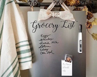 May include: A silver magnetic grocery list board with a handwritten grocery list, a white dry erase marker, and a small sticky note with a handwritten message. The board is hanging on a metal bar with decorative knobs.