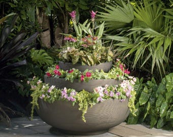 May include: A tiered, brown, outdoor planter filled with colorful flowers and greenery. The planter has a textured surface and is surrounded by lush foliage, creating a vibrant garden display. The flowers include pink and red blooms.