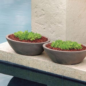 May include: Two gray, oval-shaped planters filled with green plants and reddish-brown mulch. The planters have a textured, ribbed design and sit on a concrete ledge. The background includes a pool and a textured wall.