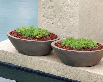 May include: Two gray, oval-shaped planters filled with green plants and reddish-brown mulch. The planters have a textured, ribbed design and sit on a concrete ledge. The background includes a pool and a textured wall.