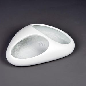 May include: A white, oval-shaped decorative bowl with two connected compartments. The interior of each compartment has a textured, frosted appearance. The letters "DG" are visible in one compartment. The bowl has a smooth, glossy exterior.