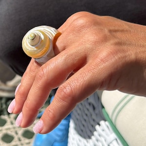 Pu&ograve; includere: Un anello a forma di conchiglia a spirale, color crema con un centro giallo dorato. L'anello &egrave; indossato su un dito, evidenziando la consistenza e il design naturali. Un gioiello unico.