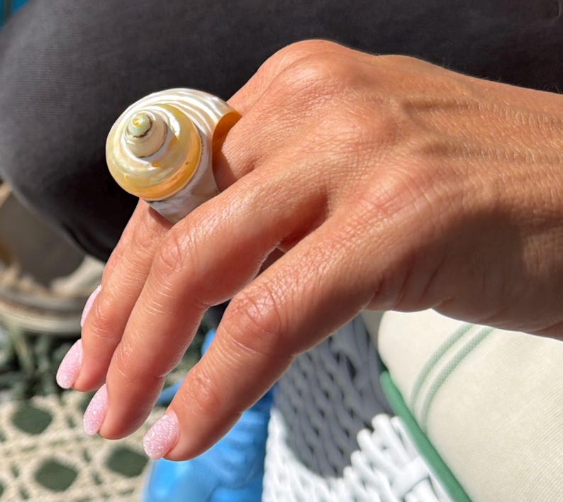 Pu&ograve; includere: Un anello a forma di conchiglia a spirale, color crema con un centro giallo dorato. L'anello &egrave; indossato su un dito, evidenziando la consistenza e il design naturali. Un gioiello unico.