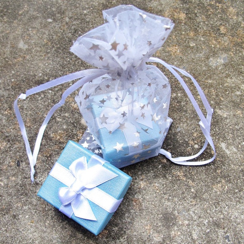 Pu&ograve; includere: Due piccole scatole regalo azzurre con fiocchi di nastro bianchi. Una scatola &egrave; all'esterno di un sacchetto trasparente bianco con accenti di stelle argentate. La borsa &egrave; legata con un nastro bianco. Le scatole sono su una superficie grigia strutturata.