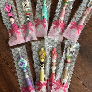 May include: A collection of decorative box cutters, each packaged in a clear bag with a pink bow and the words "For You." The cutters feature colorful beaded handles and various decorative toppers, including a diamond, rooster, and dog.