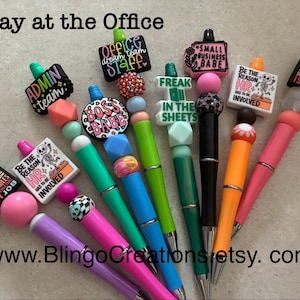 May include: A collection of colorful pens with decorative tops. The pens feature various designs and phrases, including "ADMIN team," "Boss Babe," and "HR." The pens are in shades of pink, purple, green, blue, and orange.