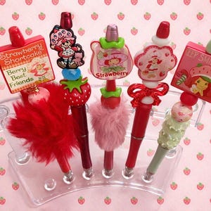 May include: A selection of decorative pens with red barrels and diverse toppers. The toppers feature a red strawberry, a pink cloud with a character, and a pink sign that says "Keep Smiling". The pens are displayed on a clear plastic stand.