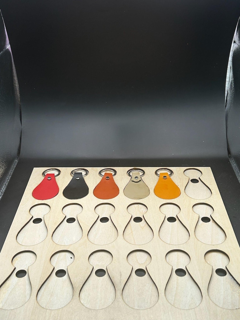 May include: A wooden board with 18 cutouts shaped like teardrop keychains. Five keychains are shown in the top row, in red, black, orange, beige, and orange. The board is made of light wood.
