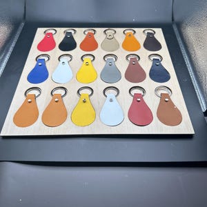 May include: A wooden board with 18 leather keychains in various colors, including red, orange, yellow, green, blue, brown, black, and white. The keychains are arranged in a grid pattern.