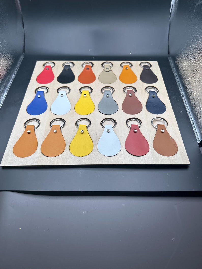 May include: A wooden board with 18 leather keychains in various colors, including red, orange, yellow, green, blue, brown, black, and white. The keychains are arranged in a grid pattern.