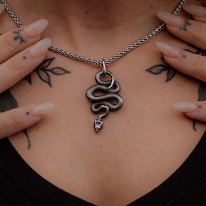 Könnte beinhalten: Ein silberfarbener Schlangenanhänger an einer passenden Metallkette. Der Anhänger zeigt ein gewundenes Schlangendesign mit detailliertem Kopf und strukturiertem Körper, das mittig über einem schwarzen V-Ausschnitt-Oberteil hängt.