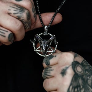 Op de afbeelding: Zilverkleurige hangerketting met een pentagramontwerp en een geitenkop in het midden. De ketting heeft een donkere ketting en wordt tegen een zwarte achtergrond gehouden. De hanger is gedetailleerd en ingewikkeld.