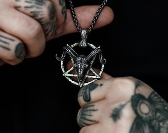 Op de afbeelding: Zilverkleurige hangerketting met een pentagramontwerp en een geitenkop in het midden. De ketting heeft een donkere ketting en wordt tegen een zwarte achtergrond gehouden. De hanger is gedetailleerd en ingewikkeld.