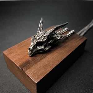 Puede incluir: Un collar con un colgante de calavera de dragón plateado en una cadena plateada. El colgante está detallado con escamas y cuernos texturizados. El collar se muestra en un bloque de madera.