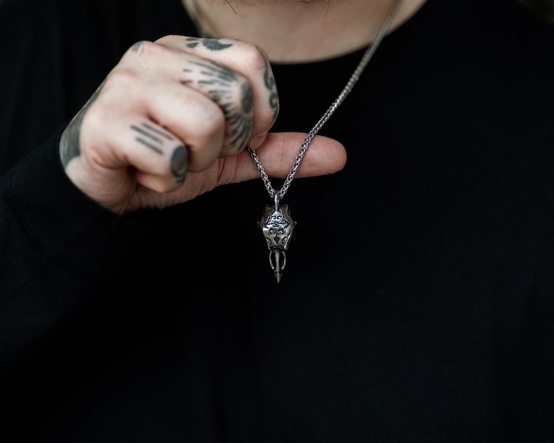 Pode incluir: Um colar com pingente de prata com um design detalhado e ornamentado. O pingente &eacute; suspenso por uma corrente delicada. O colar &eacute; exibido em um fundo preto.