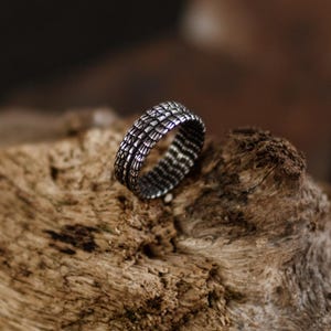 Peut inclure: Une bague argentée texturée avec un motif répétitif de formes rectangulaires. La bague est mise en valeur sur un fond en bois flou, soulignant le design complexe et l'éclat métallique du bijou.