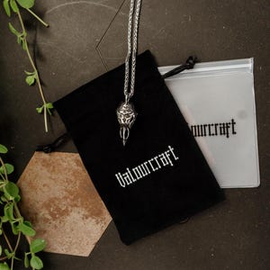 Pode incluir: Um colar com pingente prateado com um design ornamentado &eacute; exibido sobre uma bolsa preta com cord&atilde;o. A bolsa tem a palavra "Valmurcraft" impressa em branco. Uma bolsa de pl&aacute;stico transparente com o mesmo texto tamb&eacute;m &eacute; vis&iacute;vel. Folhagem verde e uma telha hexagonal marrom no fundo.