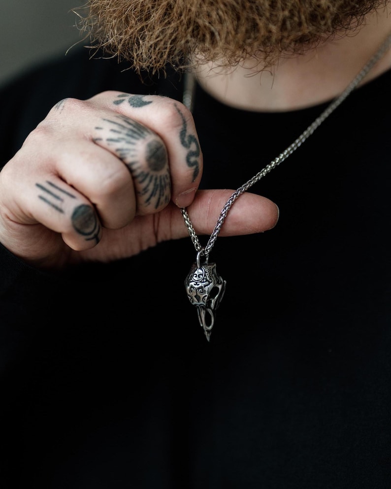 Pode incluir: Um colar com pingente prateado com um design de caveira ornamentado. O pingente &eacute; suspenso por uma corrente delicada. A caveira apresenta detalhes intrincados e um acabamento escuro oxidado, criando uma est&eacute;tica g&oacute;tica ou motard. O colar &eacute; exibido em um fundo escuro.