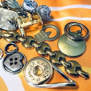 Peut inclure: Un ensemble de breloques et de pendentifs dorés, dont un breloque en forme de cœur avec un détail de bouton, un breloque rond avec le mot "Ginger" et un motif d'étoile, un breloque avec les mots "Anne Klein" et une couronne, et un breloque rond avec le mot "Coach".