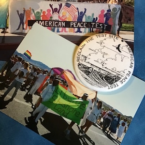 May include: A white ceramic button with black text and a graphic of a landscape. The text reads "Reclaim the Test Site American Peace Test".