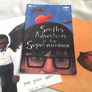 May include: Three comic books with colorful illustrations. The first book features a black and white illustration of a person wearing a white shirt and a black bow tie. The second book features a colorful illustration of a person wearing glasses and a red nose. The third book features a colorful illustration of a fish with a red and orange body.