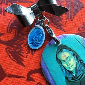 Può includere: Spilla argentata a forma di fiocco, con un ciondolo ovale blu e un ciondolo rotondo con un ritratto. La spilla è posta su uno sfondo rosso a motivi.