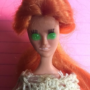 Könnte beinhalten: Eine Vintage-Puppe mit leuchtend orangefarbenem Haar und grünen Augen. Die Puppe trägt ein cremefarbenes Häkeltop. Das Gesicht der Puppe ist blass mit bemalten Gesichtszügen.