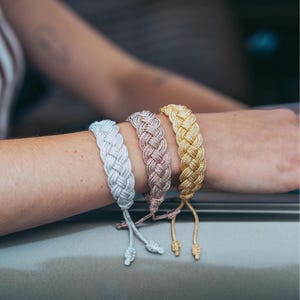 Puede incluir: Tres pulseras trenzadas en plata, oro rosa y oro. Las pulseras están atadas con un nudo en el extremo.
